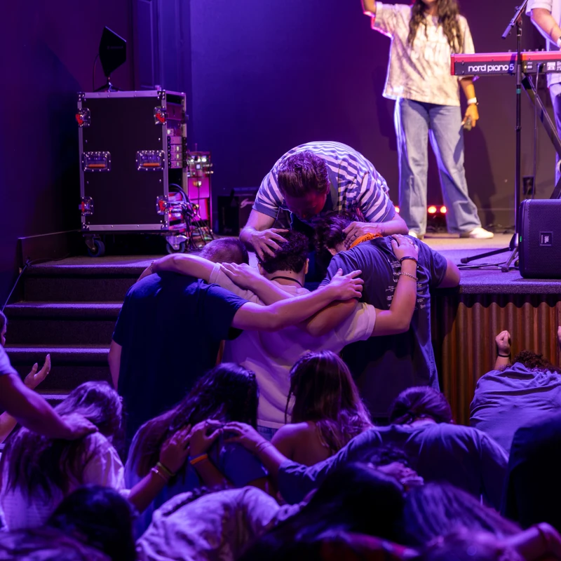 A group on a purple-lit stage huddles in a prayer circle, surrounded by an audience and a band with musical equipment.
