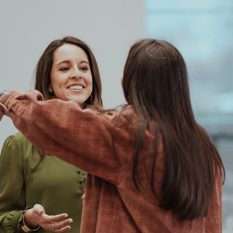 A smiling woman in a green blouse opens her arms to hug another woman who has her back to the camera.