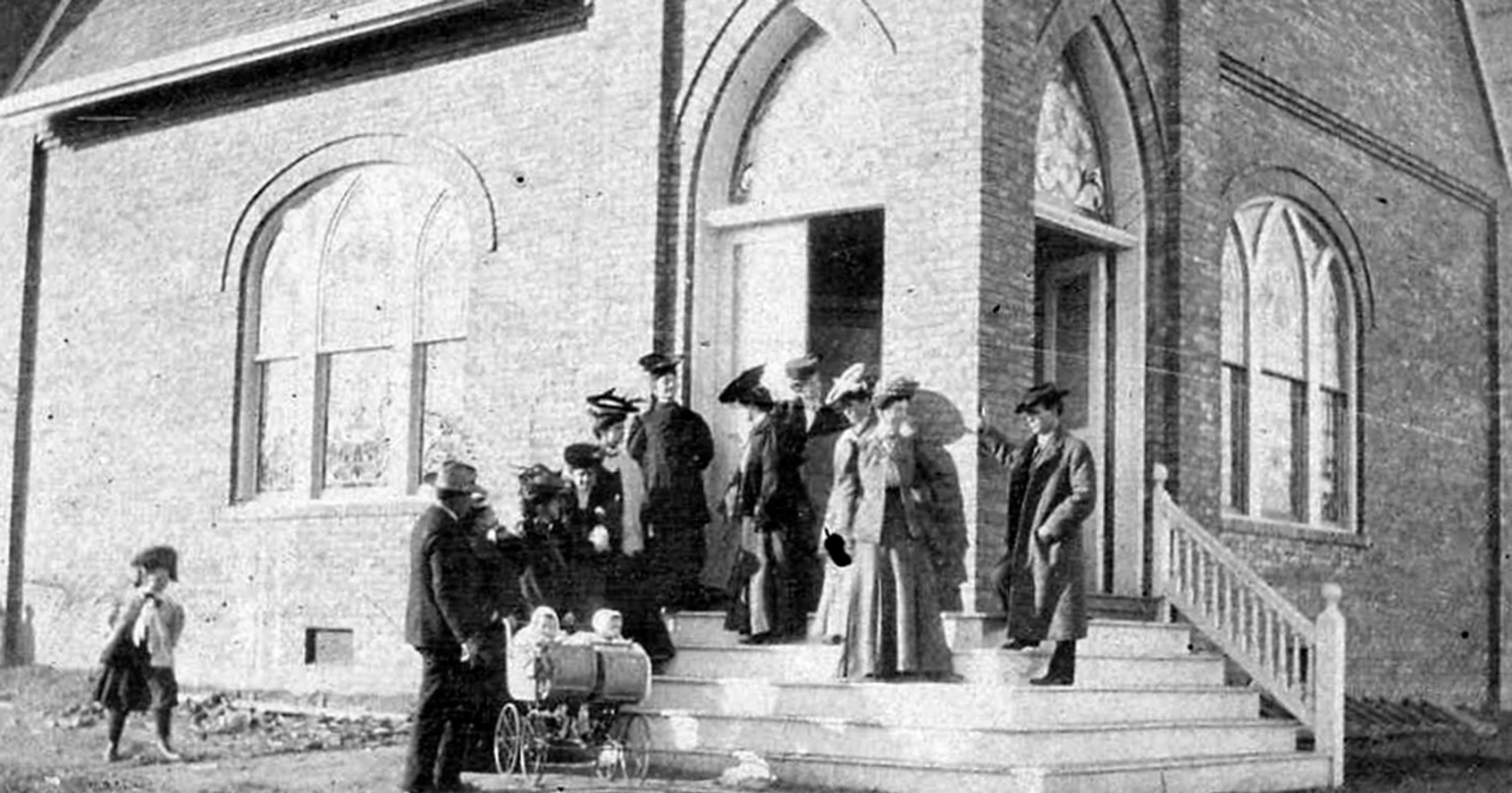 A vintage black and white photo of a congregation in period dress leaving a brick church. A man stands with a twin baby carriage at the bottom of the steps.