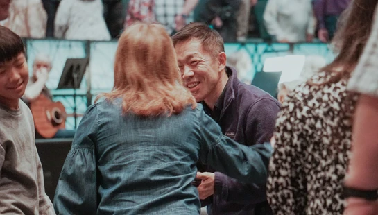 An Asian man in a purple fleece jacket smiles warmly as he talks to a woman in a denim shirt. A young boy stands to the left, also smiling.