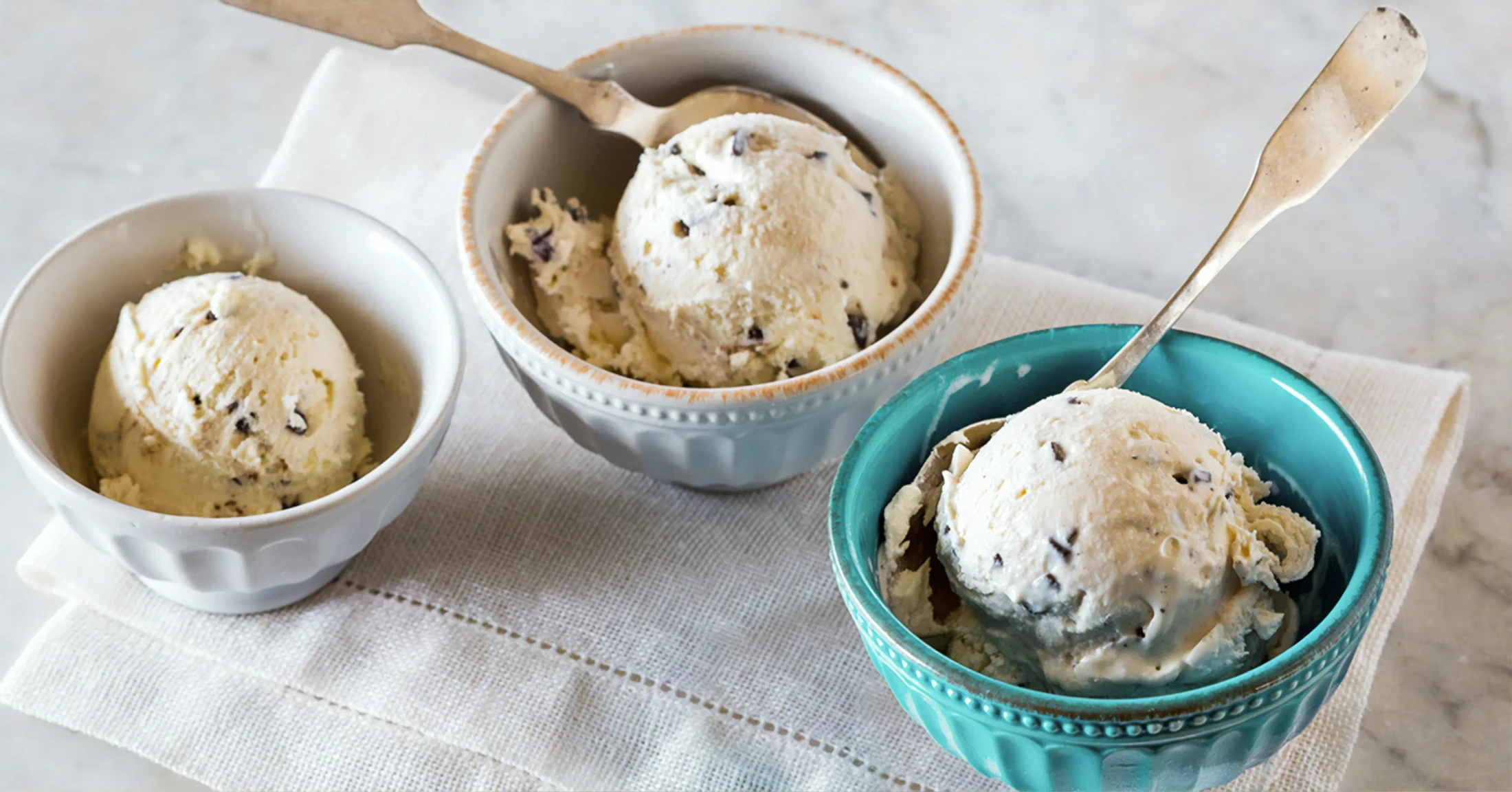 Three different ceramic bowls filled with scoops of vanilla chocolate chip ice cream, each with a spoon, sitting on a white napkin.