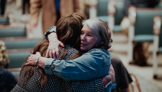 An older woman with graying hair, wearing a denim jacket, smiles gently as she warmly hugs another woman in what appears to be a church or auditorium.