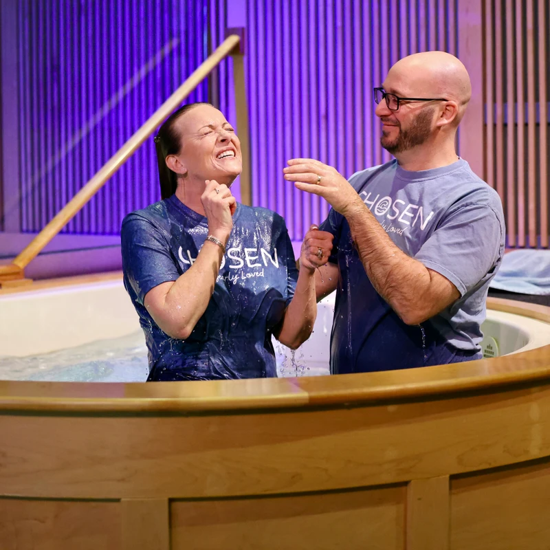 Woman celebrating in a baptismal pool after being baptized, standing beside a pastor in a church sanctuary.