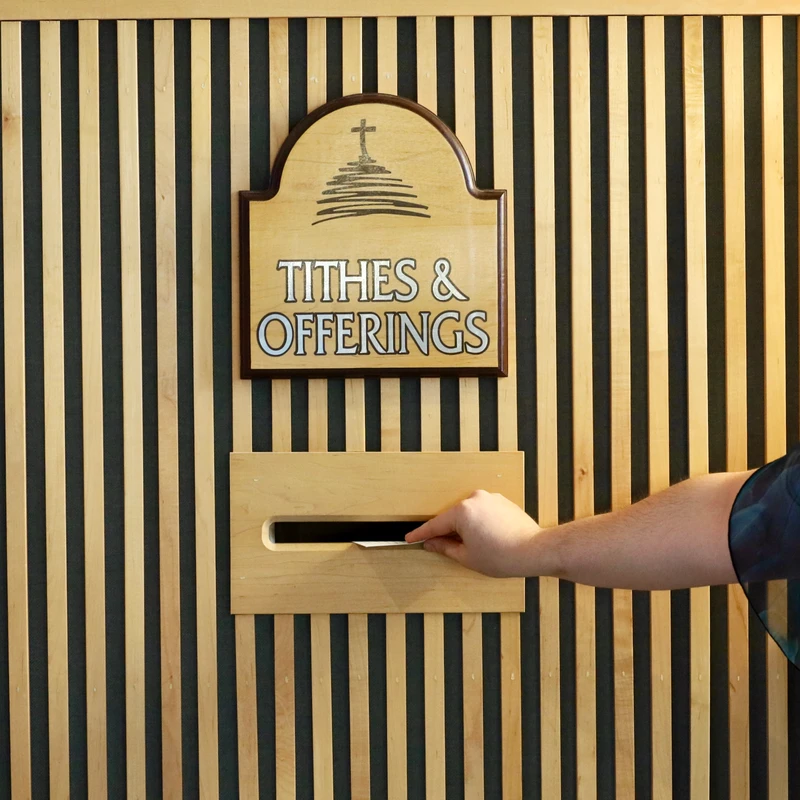 Person placing an envelope into a wooden tithes and offerings box mounted on a striped wall.