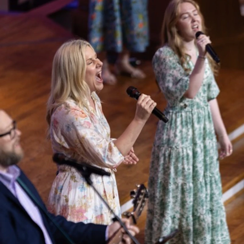 A blonde woman sings passionately into a microphone next to another woman singing. A man in the foreground plays an acoustic guitar.