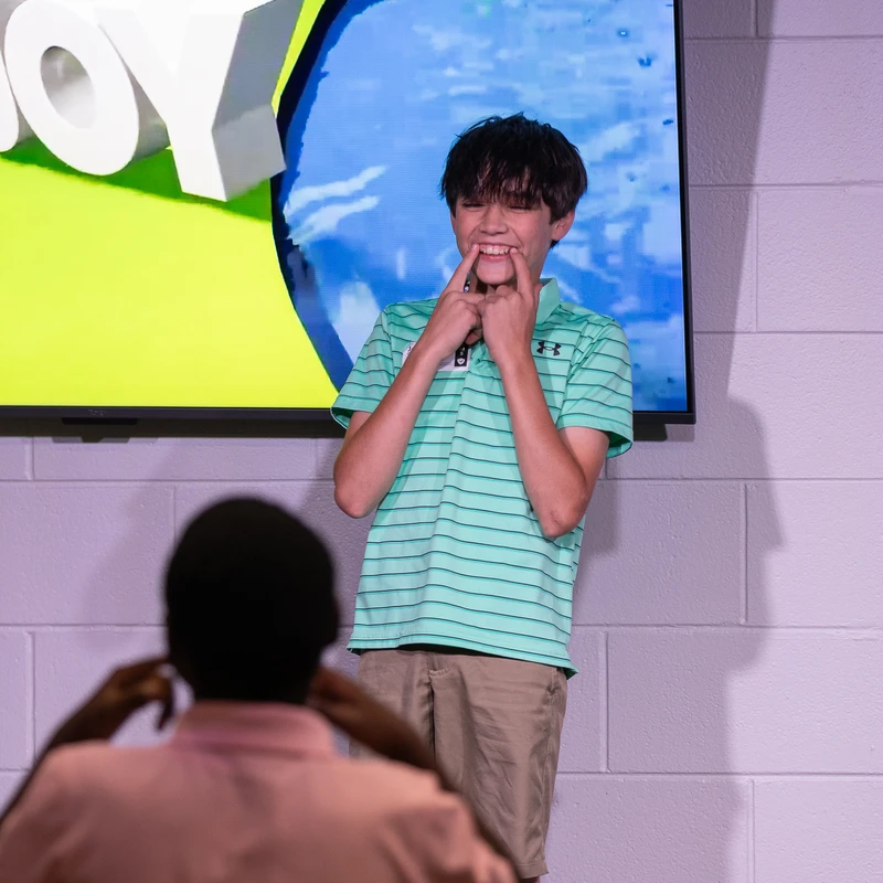 A young boy in a green striped polo shirt stands on a stage, using his fingers to make a wide, silly smile. The word 'JOY' is on a screen behind him.