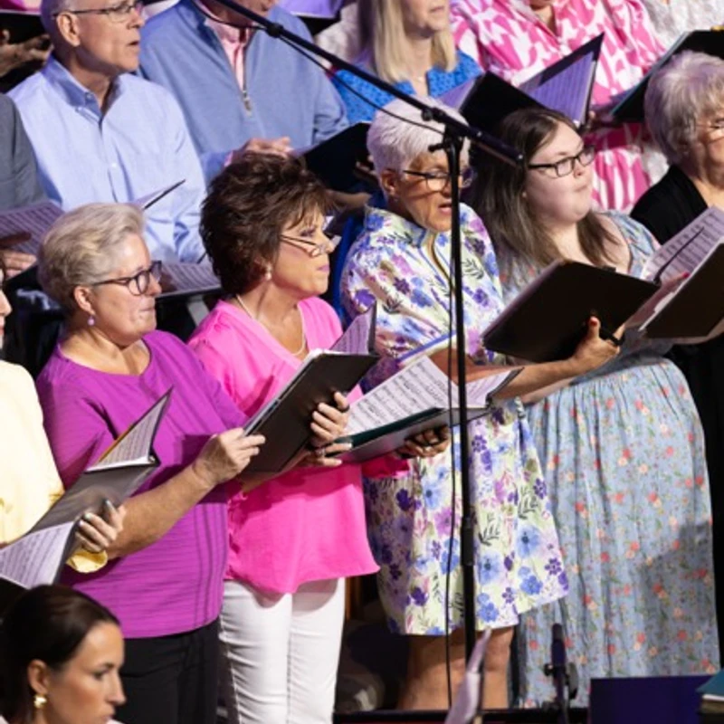 A diverse, multi-generational choir of men and women in colorful clothing sing from sheet music binders while standing on risers during a performance.