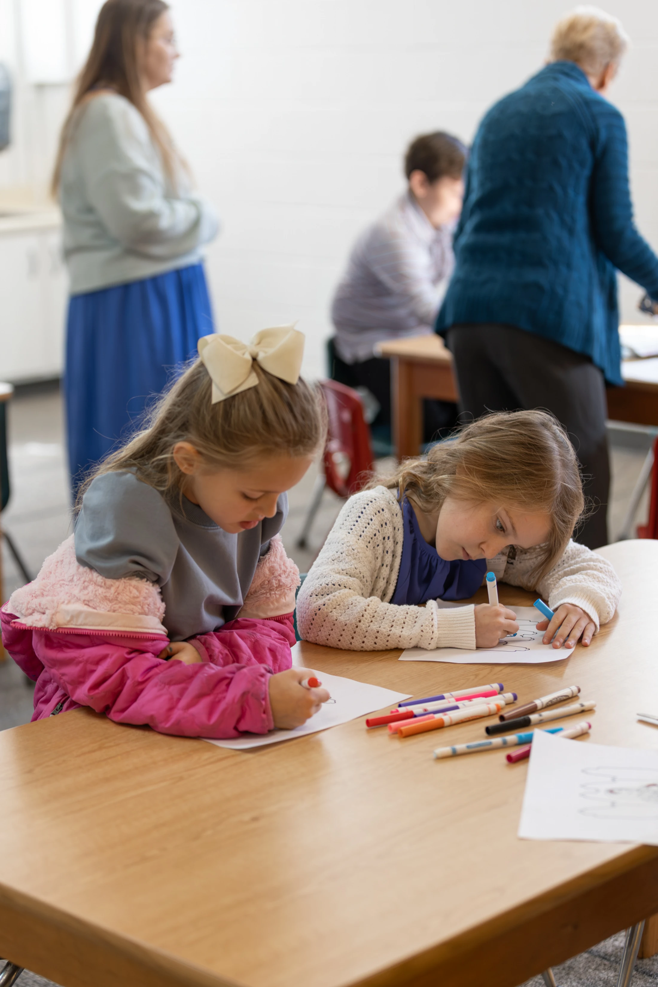 Two young girls sit side-by-side at a wooden desk, focused on coloring on paper with markers. Adults and other children are blurred in the background.