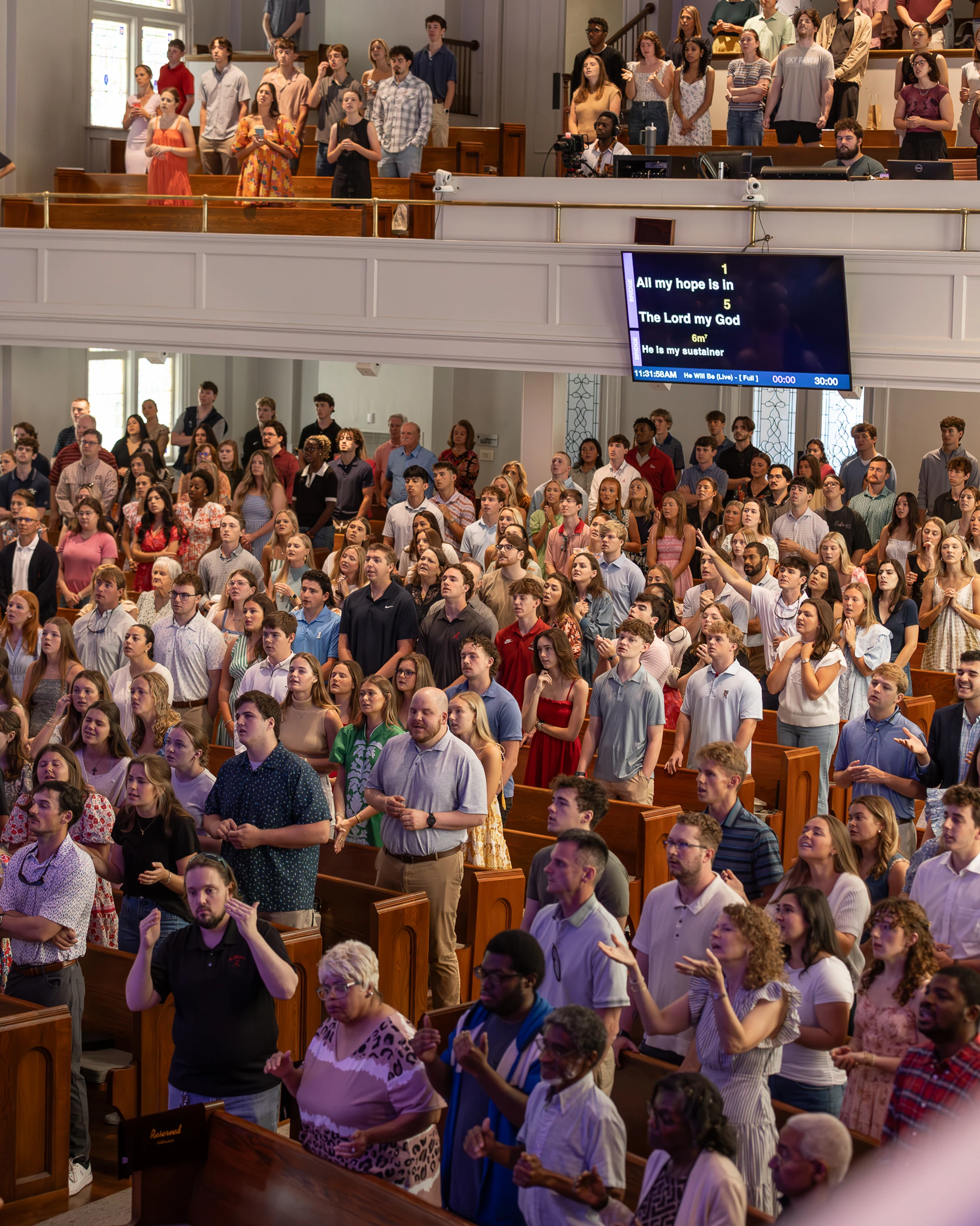 A large, diverse congregation stands in wooden pews inside a church, singing and worshiping with some raising their hands toward heaven.