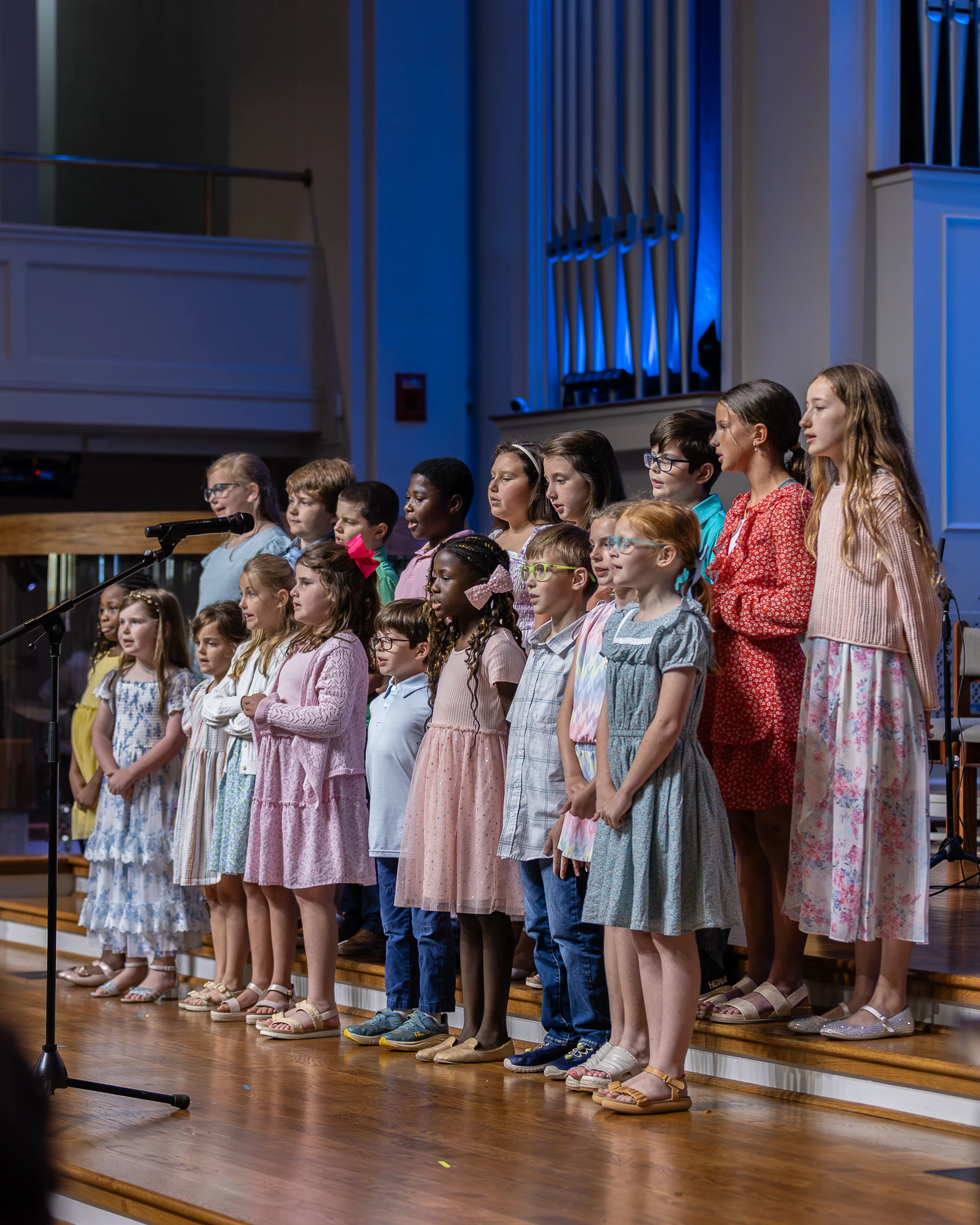 A diverse group of young children stands in rows on a wooden stage, performing as a choir. A large pipe organ lit with blue light is in the background.