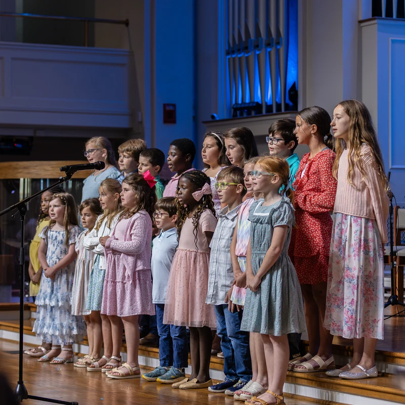 A diverse group of young children stands in rows on a wooden stage, performing as a choir. A large pipe organ lit with blue light is in the background.