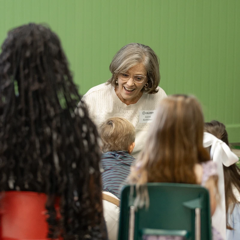 An older female teacher smiles warmly while speaking to a group of young children who are seen from behind, sitting in a classroom with a green wall.