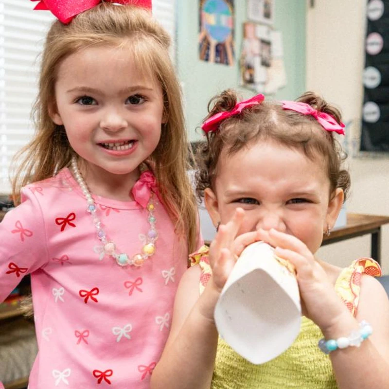 Two young girls in a classroom. One wears a pink shirt and a large pink bow and smiles. The other wears a yellow dress and playfully drinks from a paper cup.
