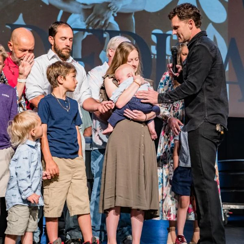 A family stands on a stage during a ceremony. A man with a microphone places his hand on an infant held by its mother as other family members look on.