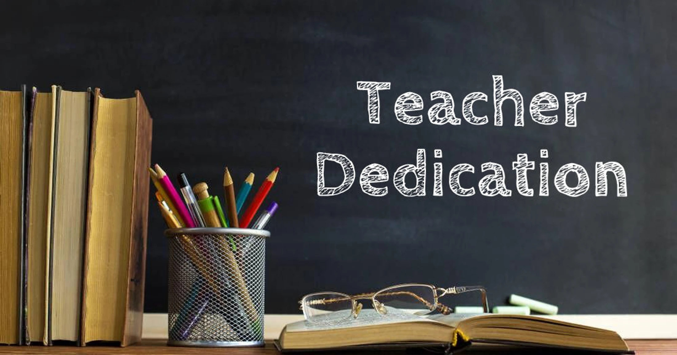 A teacher's desk with books, pencils, and glasses on a book in front of a blackboard that reads "Teacher Dedication" in chalk-style letters.