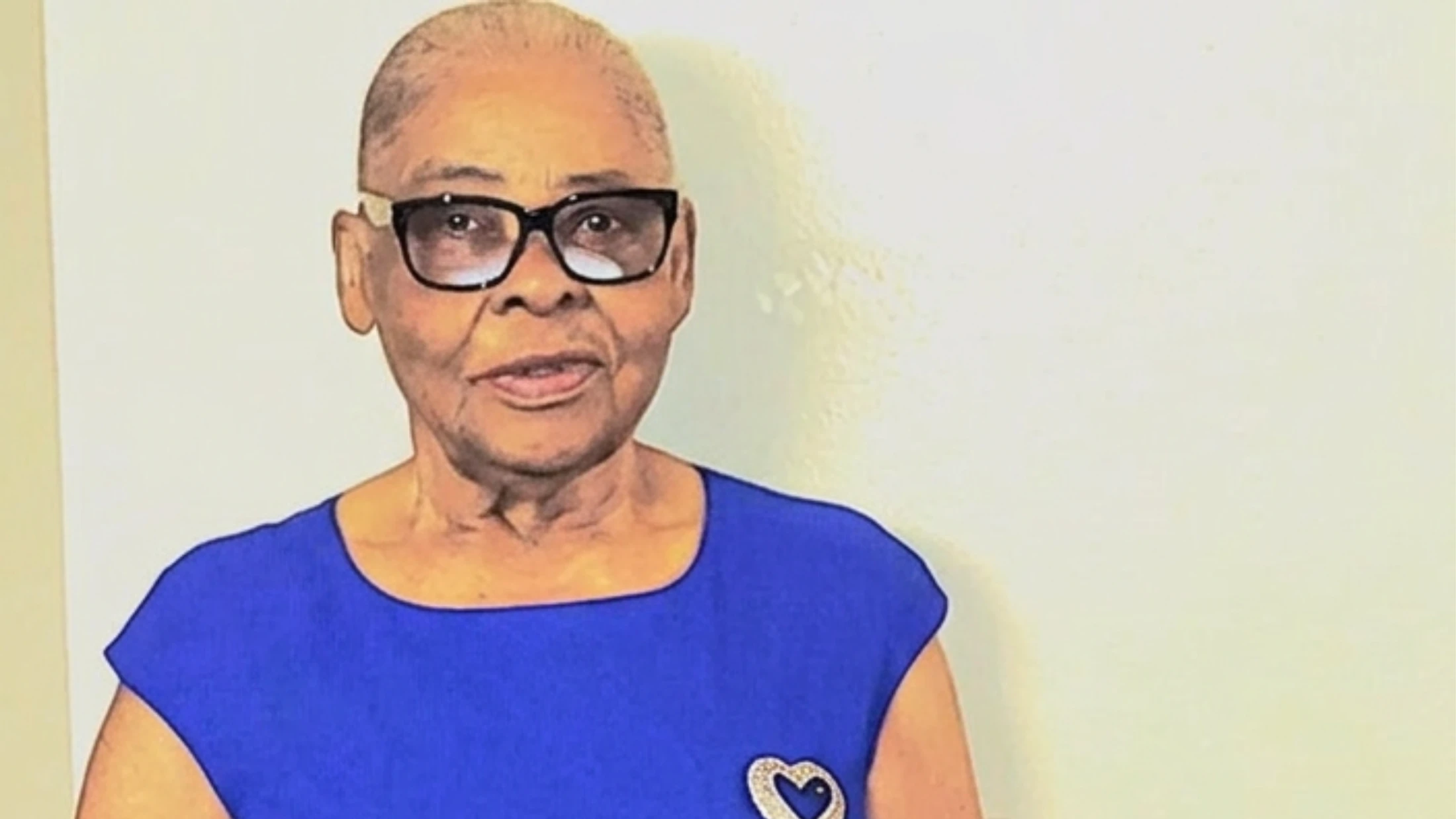 A head-and-shoulders portrait of an elderly Black woman with short hair and glasses. She wears a royal blue top with a heart brooch and looks at the camera.