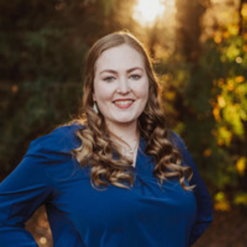 A headshot of a young woman with long, curly brown hair, wearing a blue shirt. She is smiling in front of a sunlit, leafy background.