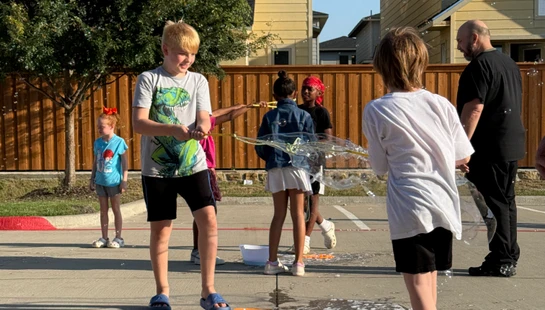 A group of children and an adult play outdoors on a sunny day, creating a giant soap bubble with a string wand and a bucket of soapy water.