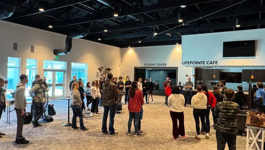 A large group of people stand in a circle in a spacious, modern hall with a high black ceiling. Signs on the wall read 'Student Center' and 'Lifepointe Cafe'.