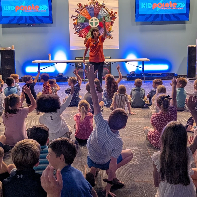 A woman on a low stage leads a group of children sitting on the floor. Many of the kids have their hands raised, enthusiastically participating in the lesson.