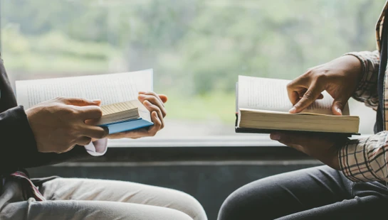 A close-up of two people sitting side-by-side, each holding an open book. One person points to a page in their book, suggesting a shared discussion.