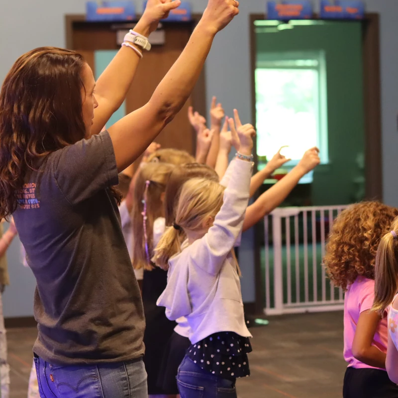 A woman with long brown hair leads a group of young girls in an activity. They all stand with their arms raised high in the air inside a large room.