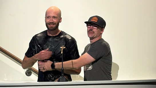 A smiling, bald man in a wet black t-shirt stands in a baptismal font after being baptized by another man in a cap and glasses standing beside him.