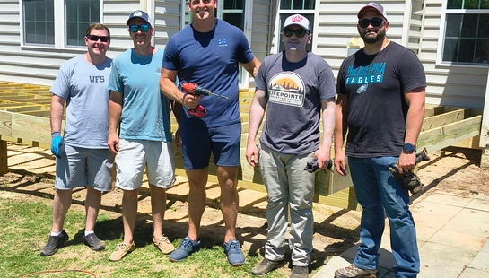 Five smiling men stand together for a photo in a backyard in front of the wooden frame of a deck they are building. One man holds a red power drill.