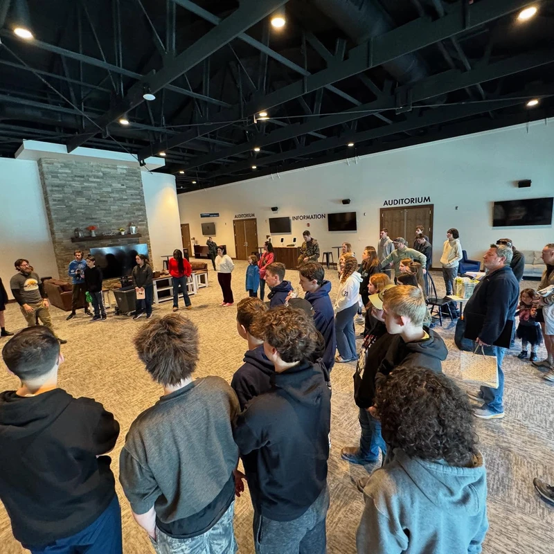 A wide-angle shot of a large group of people, mostly young adults and teens, standing in a circle in a spacious, modern lobby with a high ceiling.