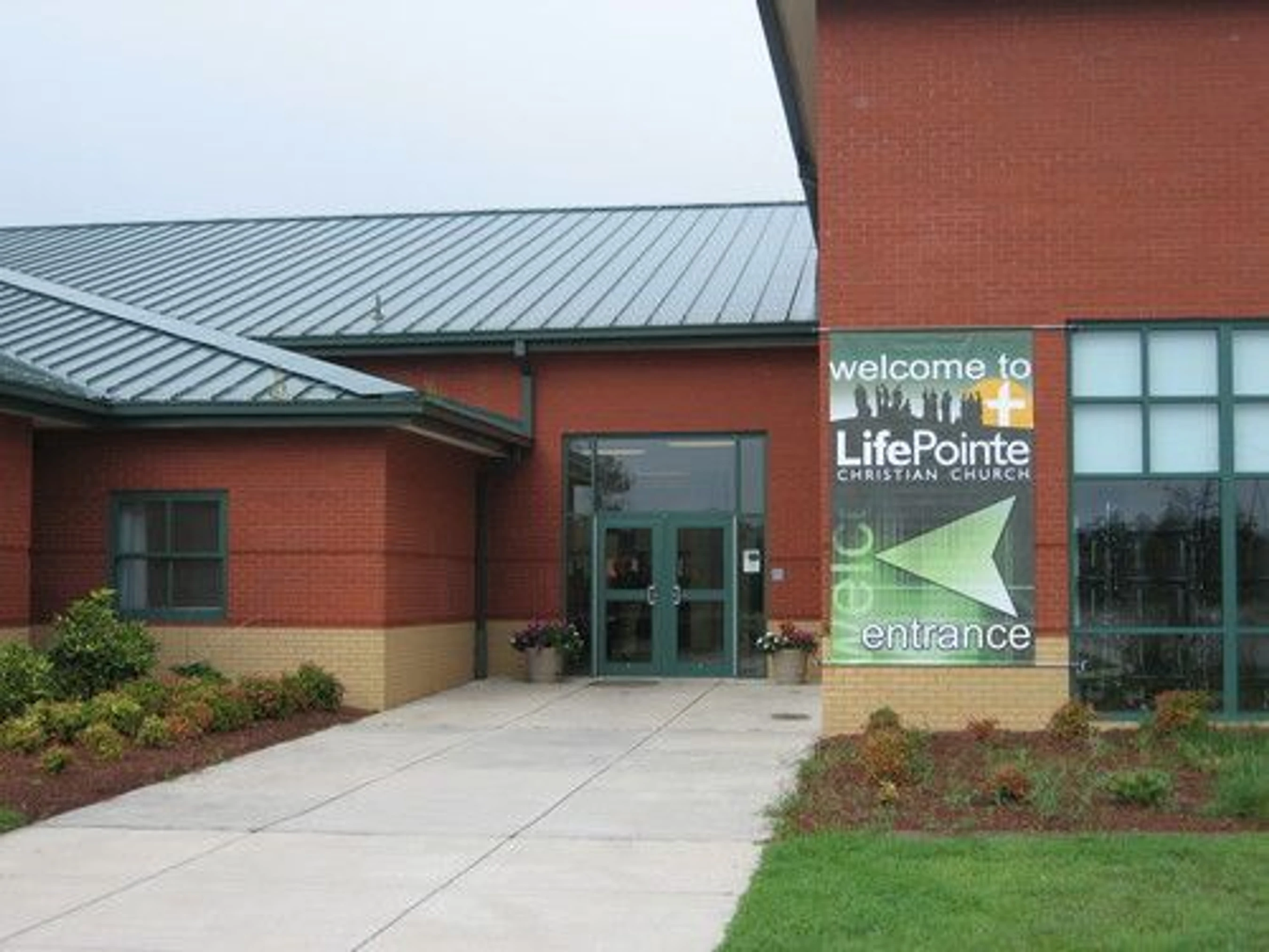 The exterior of a modern red brick church with a green metal roof. A large banner welcomes visitors to the glass double doors at the main entrance.