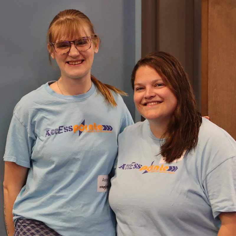 Two women stand side-by-side smiling at the camera. They are both wearing light blue t-shirts with the 'AccessPointe' logo printed on the front.