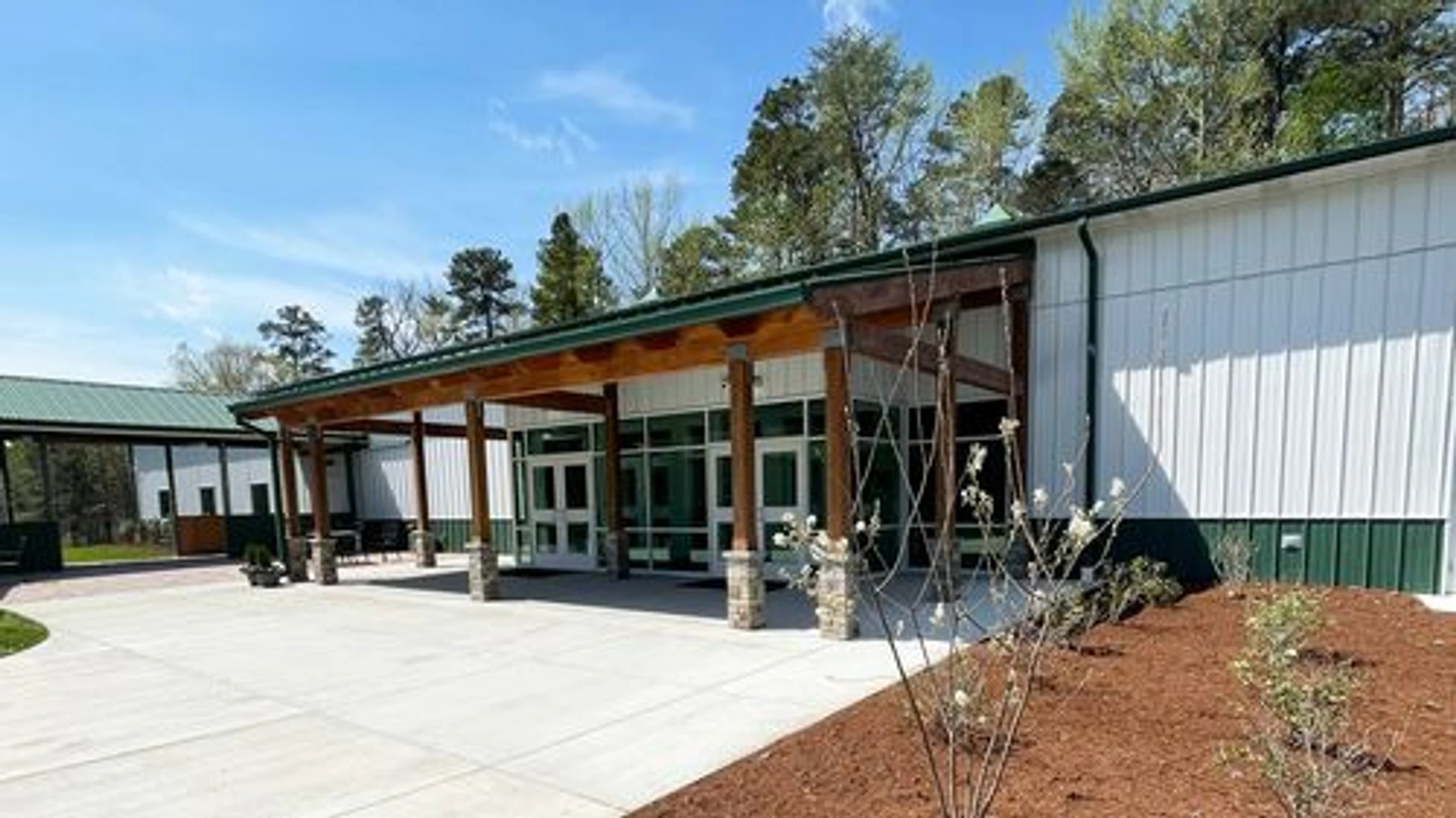 The entrance to a modern white and green building with a large wooden awning supported by stone pillars. A concrete plaza is in front under a clear blue sky.