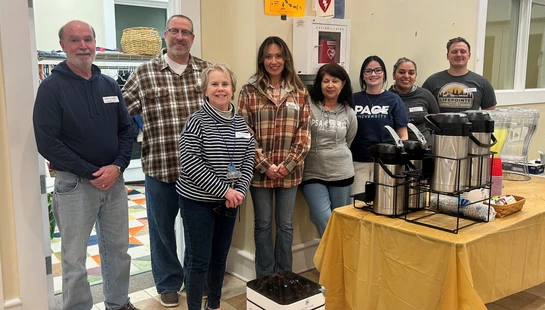 A diverse group of eight smiling volunteers standing together indoors next to a table with coffee and drinks ready for an event.