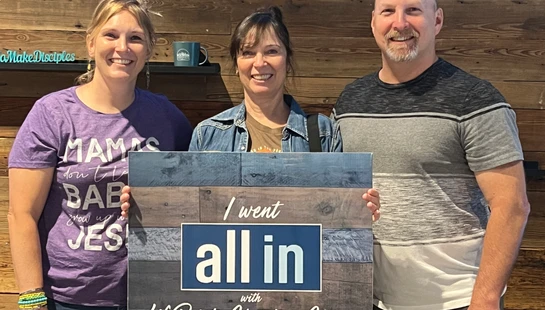 Two women and a man stand smiling in front of a wood wall, holding a sign that reads, 'I went all in with LifePointe Christian Church'.