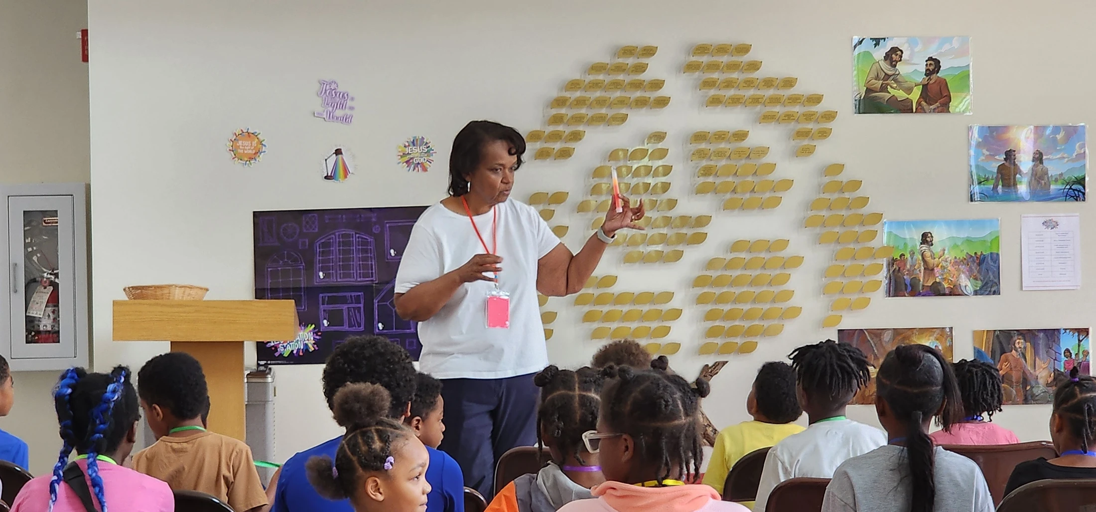 An African American woman stands and teaches a group of young children sitting in chairs, in a classroom decorated with religious pictures on the wall.