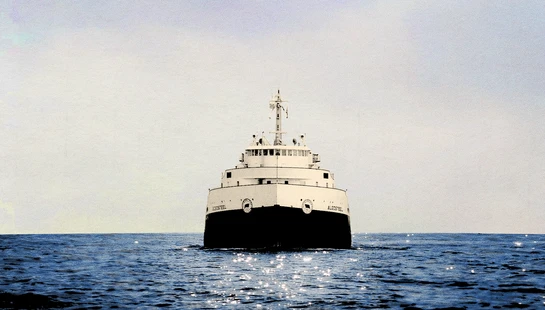 A head-on shot of a large black and white freighter, the Algosteel, sailing toward the viewer on a sparkling blue sea under a hazy white sky.