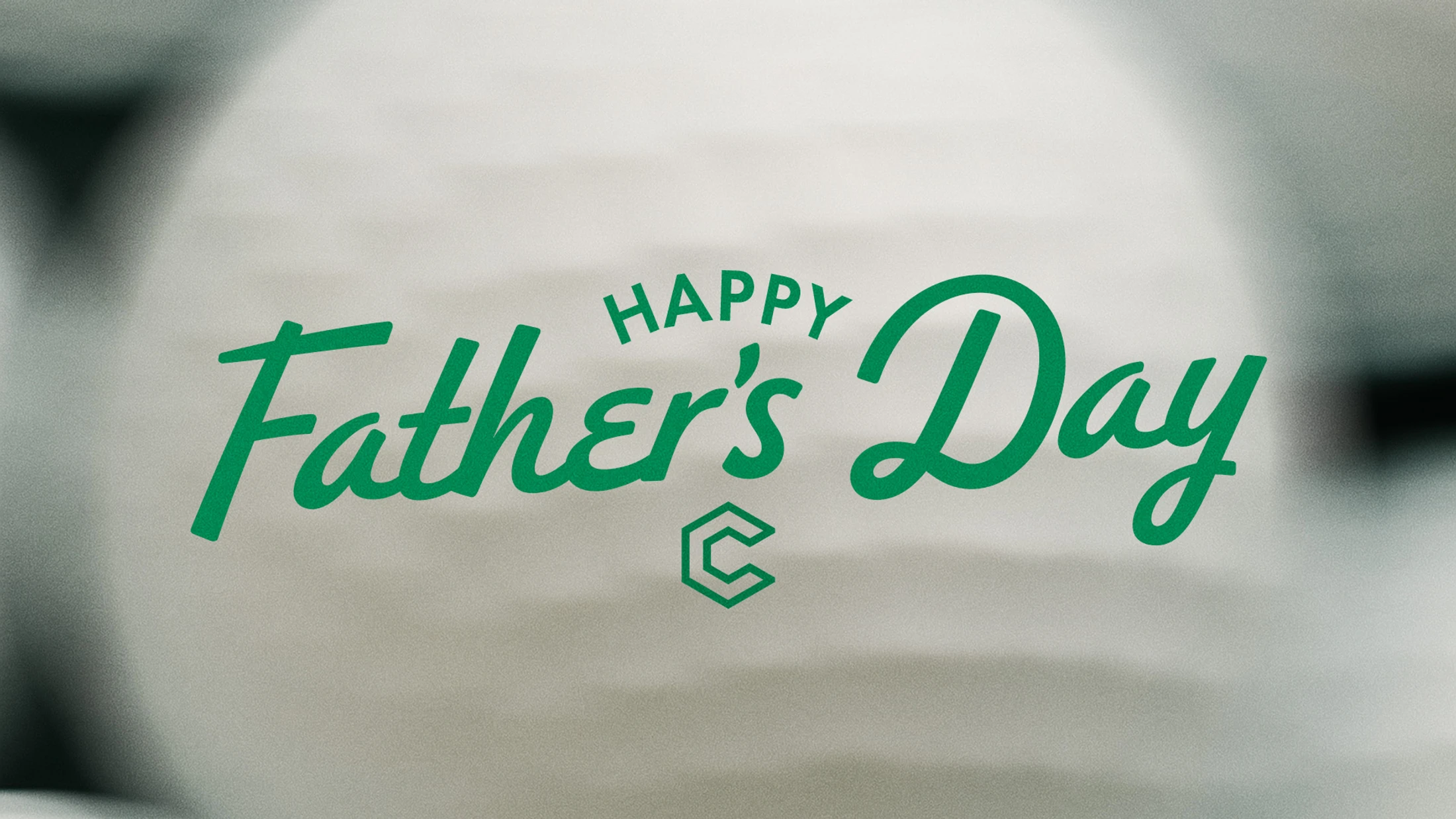 The words 'Happy Father's Day' written in a green script font over a blurry, soft-focus grey background with a small geometric logo underneath.