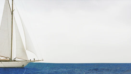 The bow of a classic white sailboat with full sails on a deep blue ocean under a bright white sky. The photo has a vintage, textured effect.