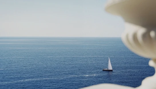 A high-angle view of a lone sailboat on the deep blue ocean, seen past the soft-focus silhouette of a stone balustrade on a sunny day.