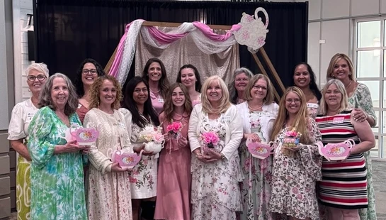 A group of thirteen women in dresses smile for a photo at a tea party, holding decorative teapots in front of a backdrop with a 'Connect' sign above.