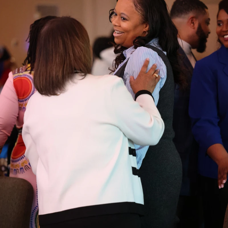 A smiling Black woman with long dark hair is embraced by another woman in a white jacket during a social gathering with other people in the background.