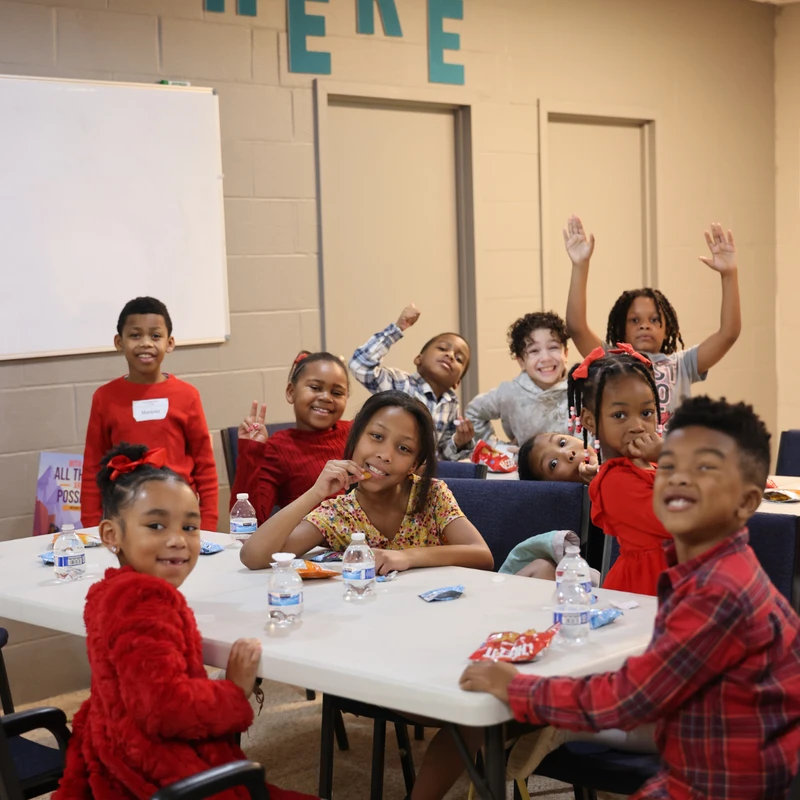 A group of happy young children sitting at a table in a classroom, smiling and waving at the camera while enjoying snacks and bottled water.