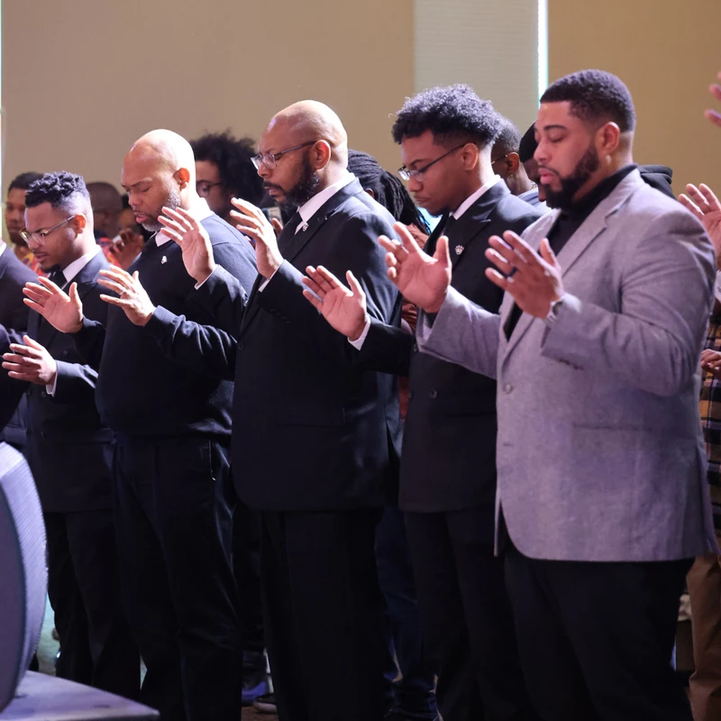 A line of Black men dressed formally stand side-by-side with their eyes closed and hands raised in prayer or worship.