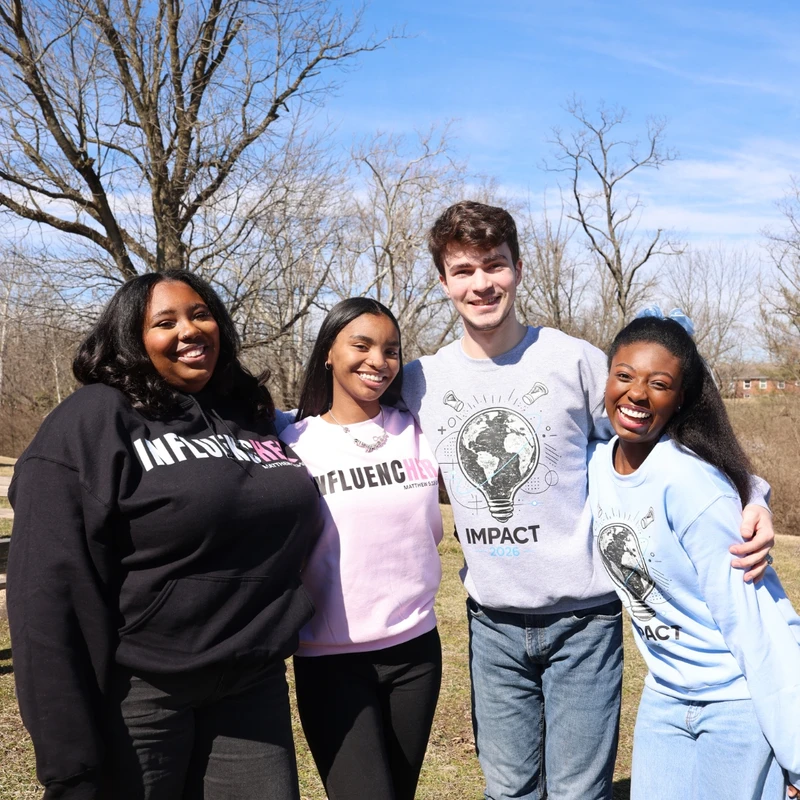 A group of four diverse young adults, three women and a man, smiling happily while posing together outdoors in a park on a sunny day with bare trees.
