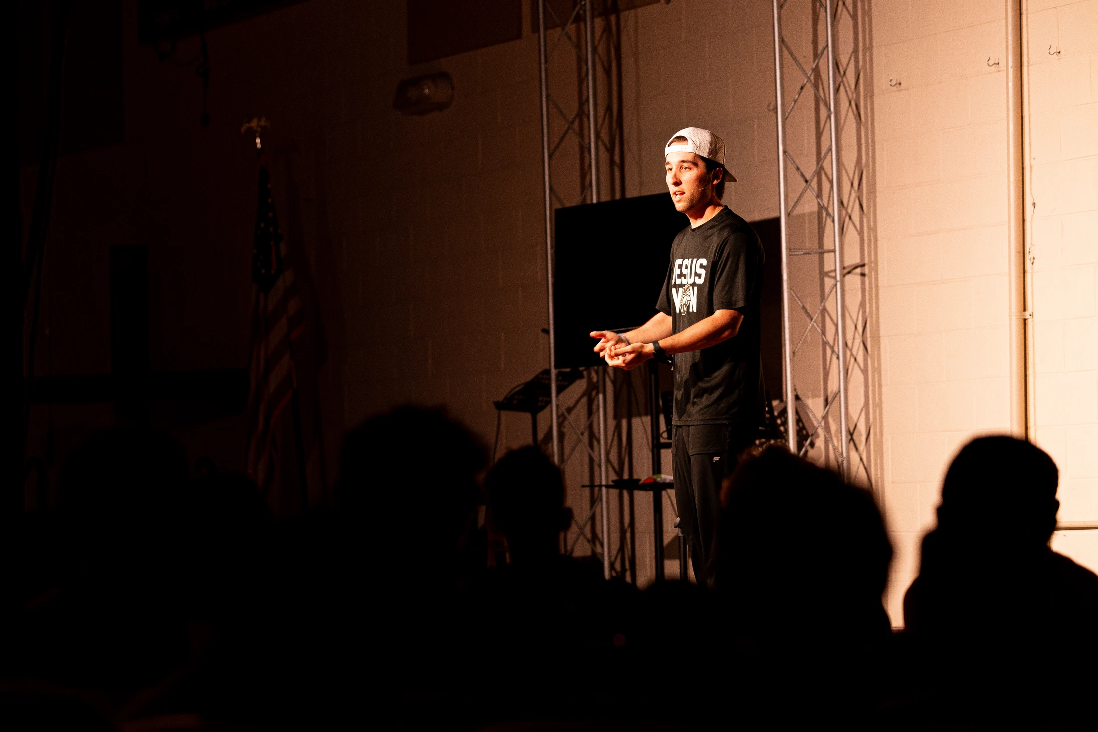 A young man in a black t-shirt and white baseball cap stands on a dimly lit stage, speaking to an audience whose silhouettes are visible in the foreground.