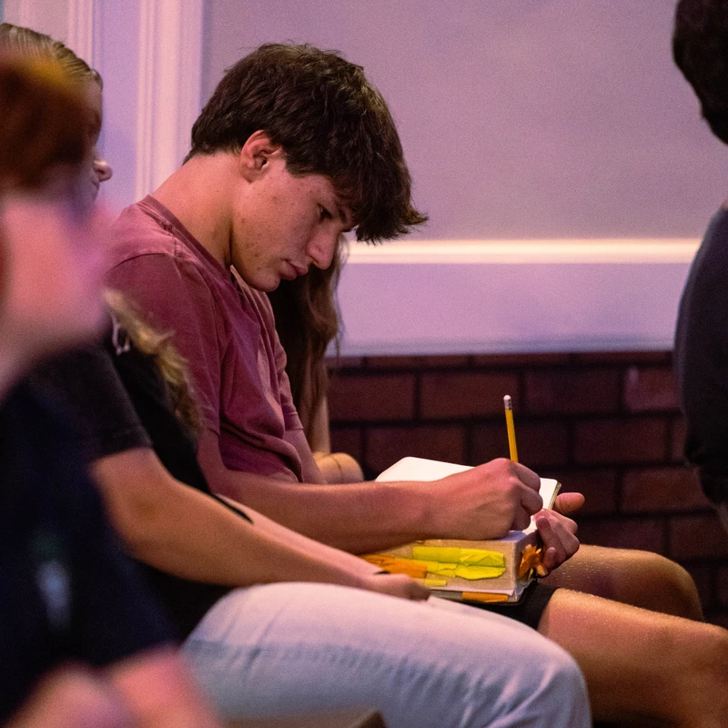 A young man with dark hair sits among other people, looking down and writing with a pencil in a notebook resting on his lap in a dimly lit room.