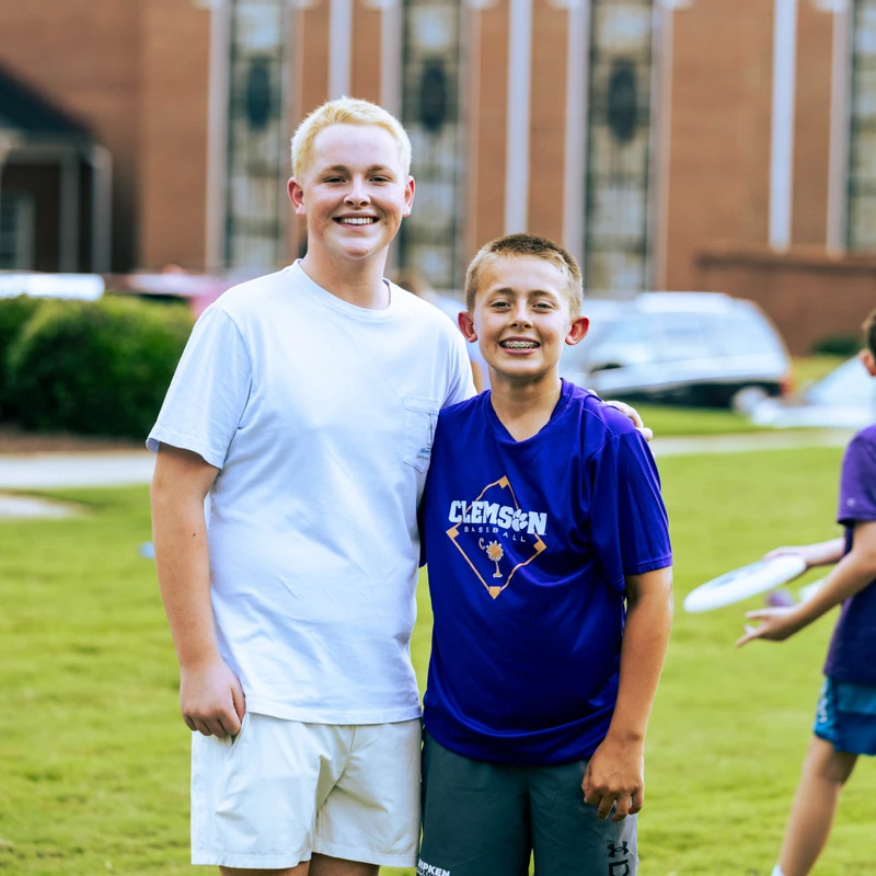 Two smiling boys, one older with blond hair and one younger with braces, stand with arms around each other on a green lawn in front of a brick building.