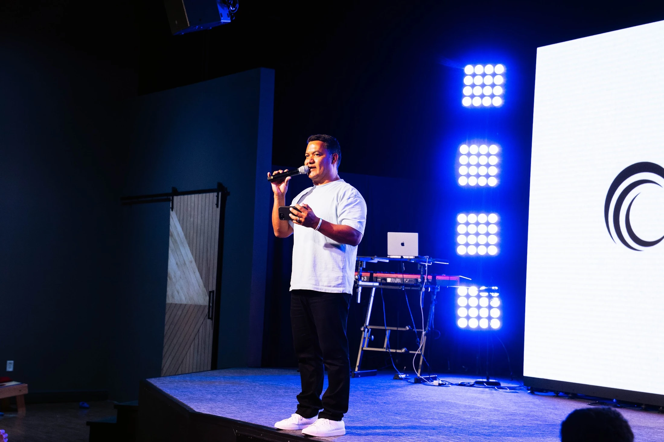 A man in a light-colored t-shirt and dark pants stands on a stage, speaking into a microphone. The stage is lit with bright blue lights.