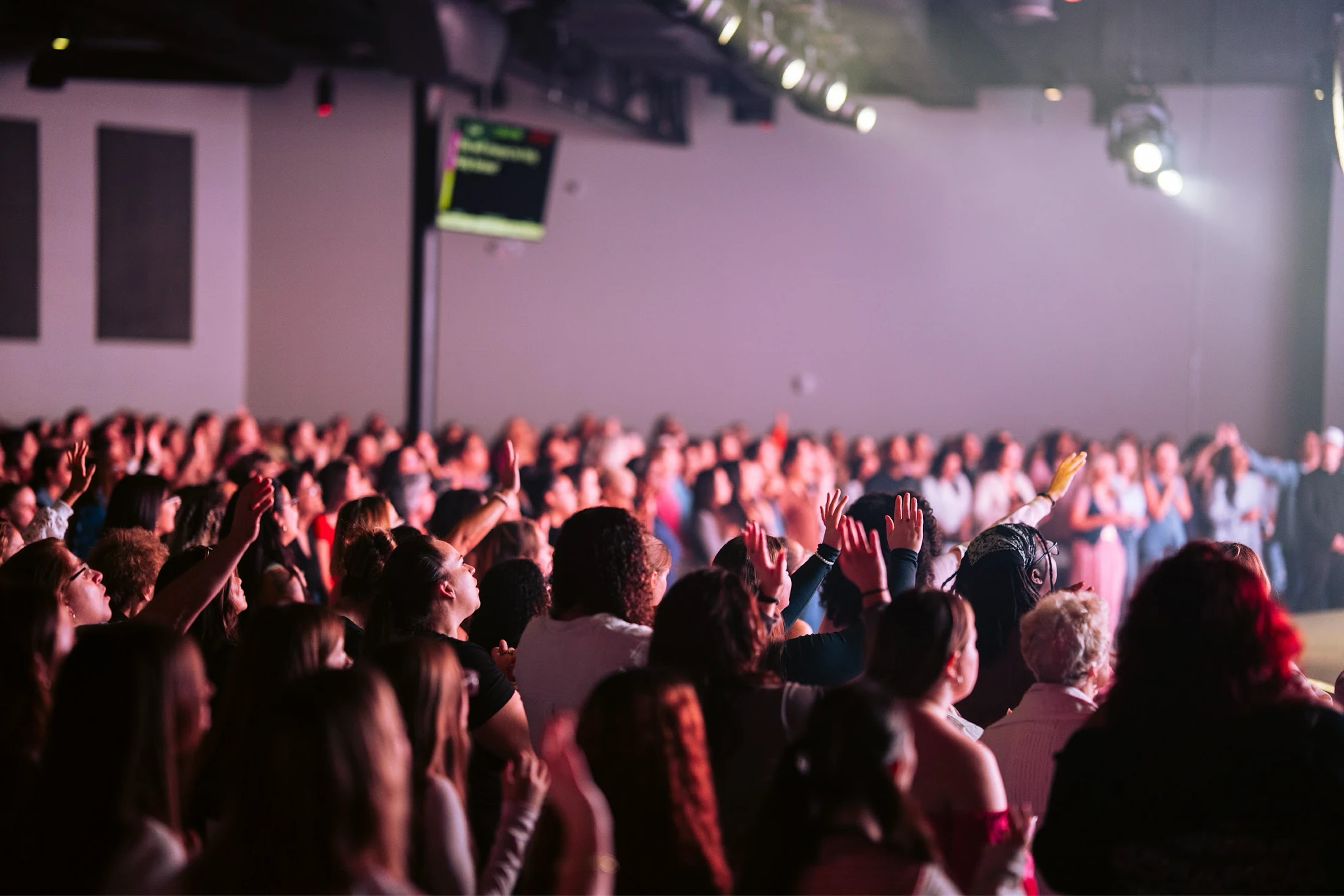 A view from behind a large crowd in an auditorium. Many people have their hands raised in the air in an act of worship or praise under stage lights.