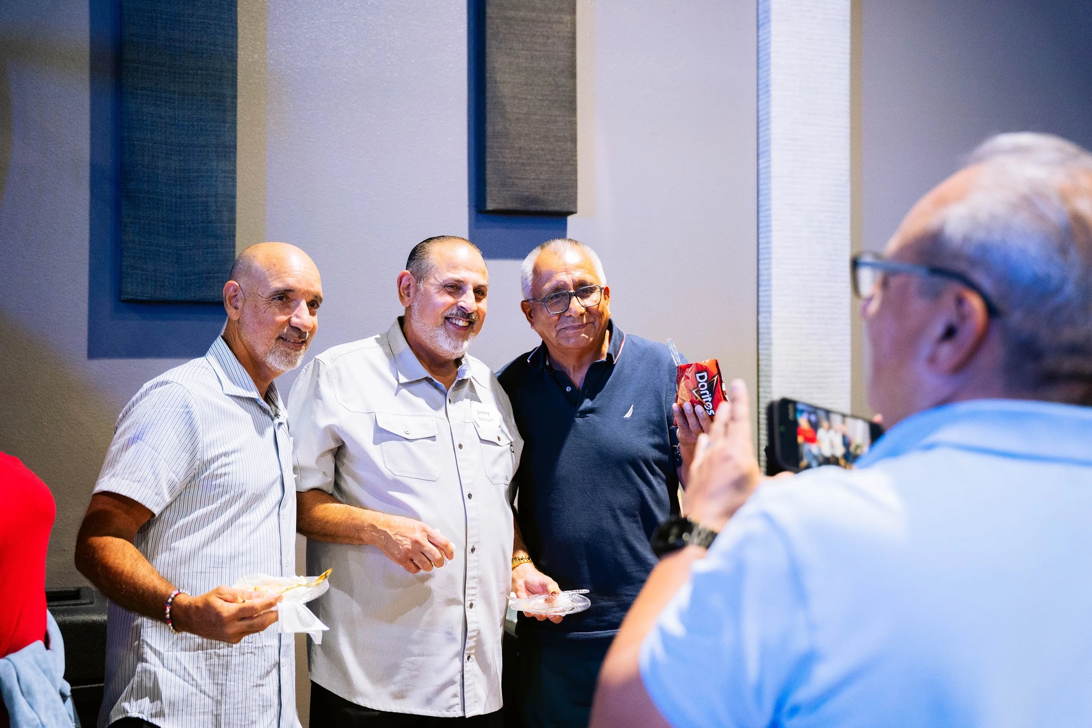 Three men stand together and smile while a fourth man, seen from behind, takes their picture with his smartphone during an indoor gathering.